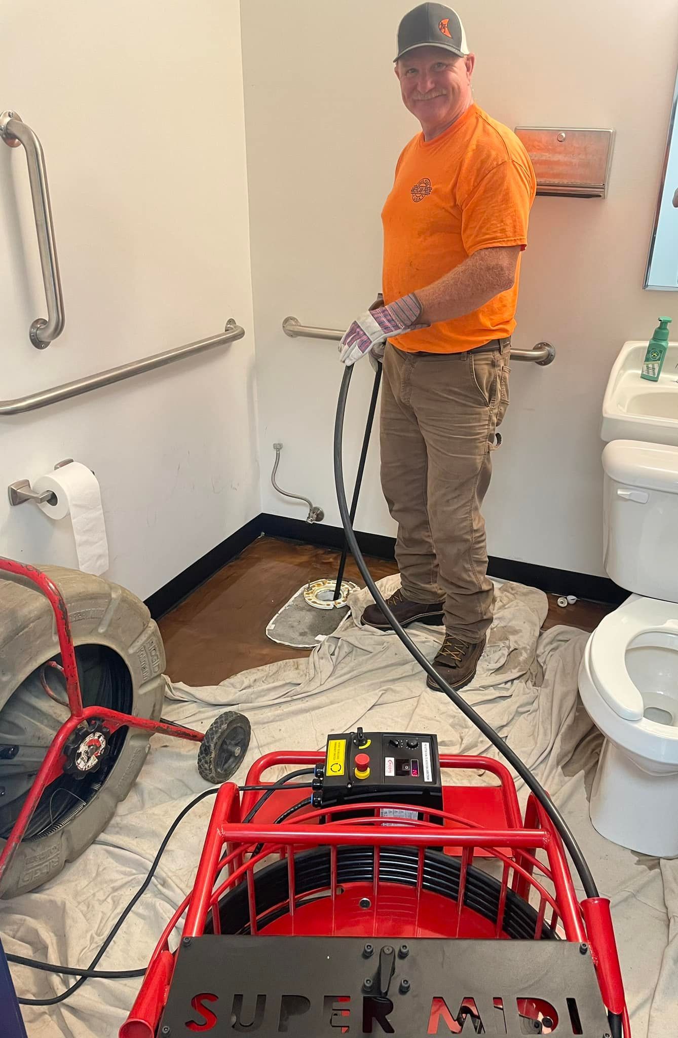 Plumber using a drain cleaning machine in a bathroom. Man in orange shirt, brown pants. Red machine on floor.