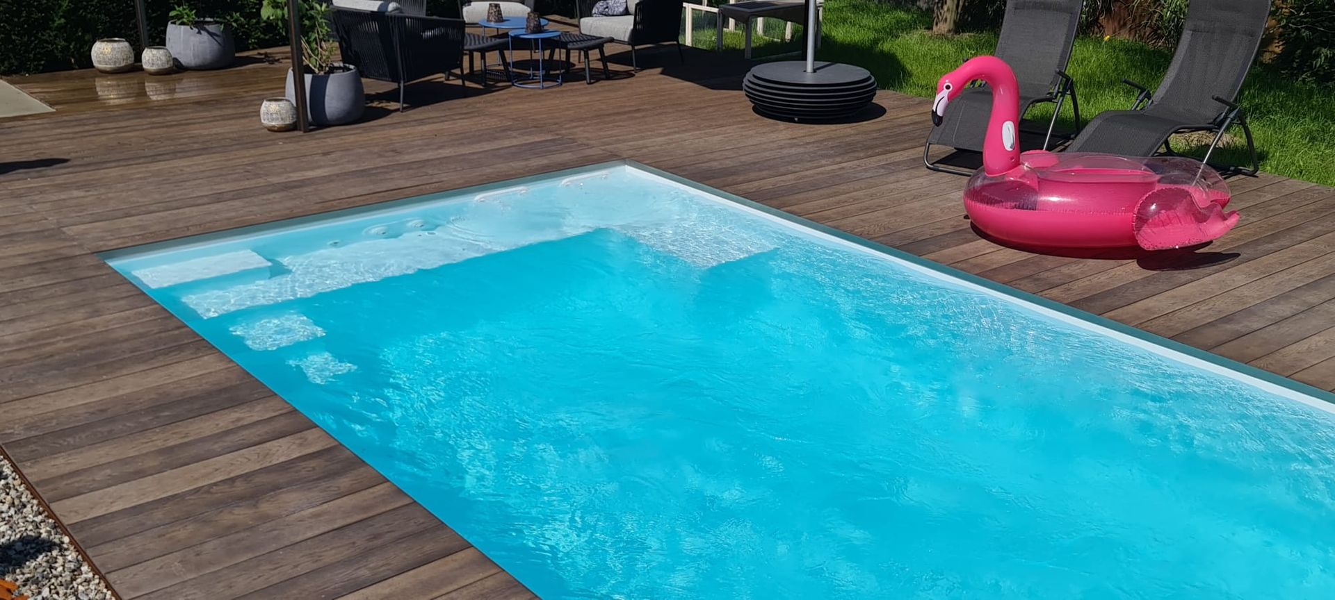 Een rechthoekig zwembad met houten vlonder, een roze flamingo-zwemband en ligstoelen.