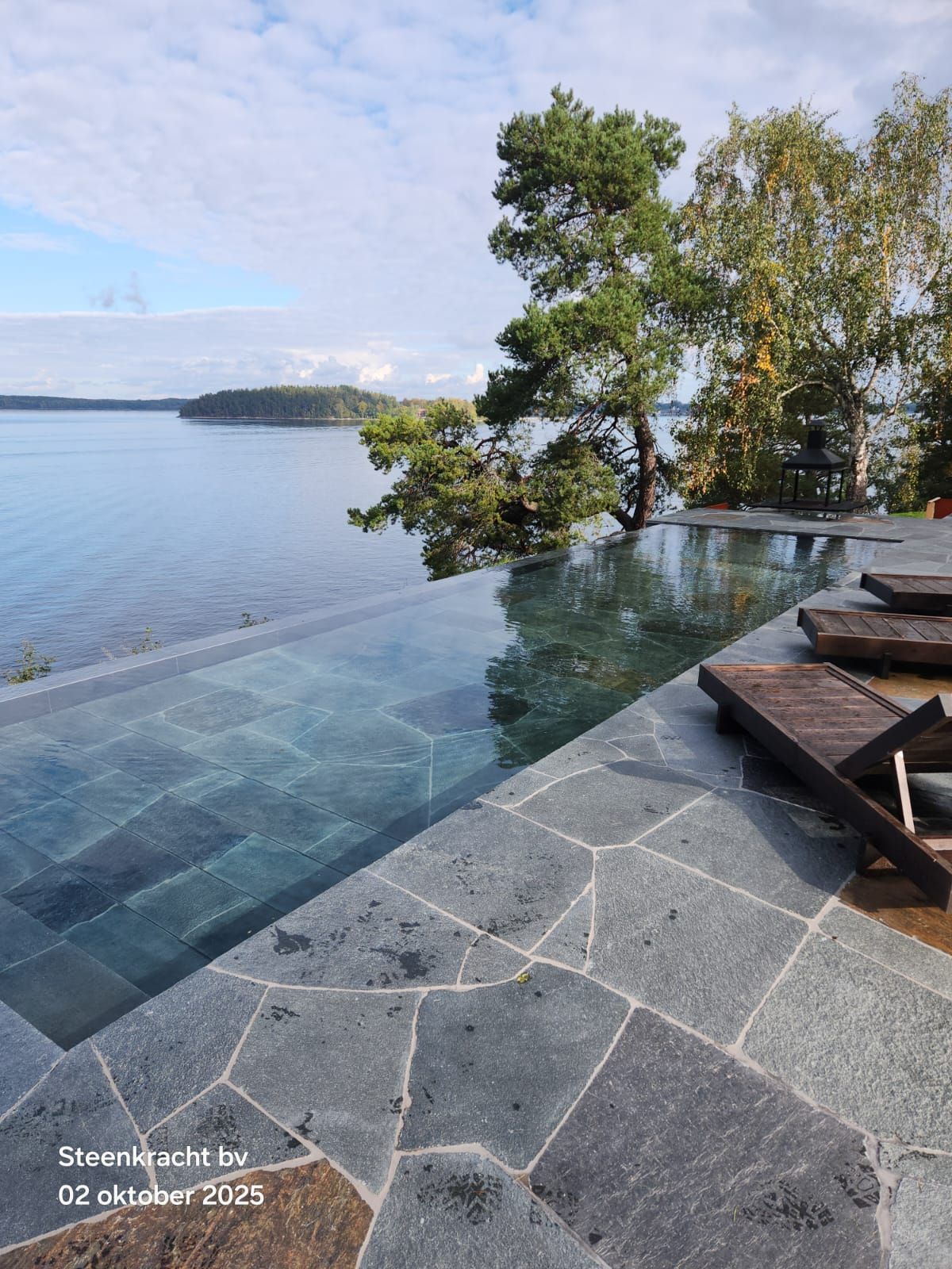 Infinity-zwembad (MagicPool) met uitzicht op een meer met bomen. Terras met leistenen tegels en ligstoelen.