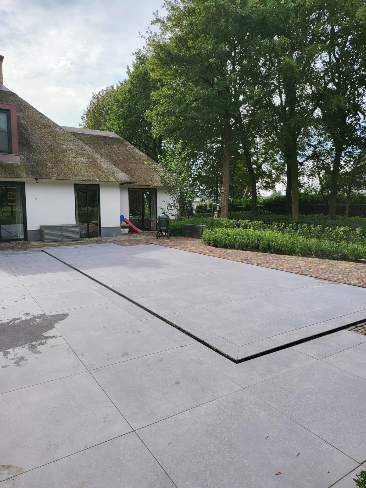 Een grijs stenen terras naast een wit huis met een rieten dak, omgeven door bomen en groen. MagicPool