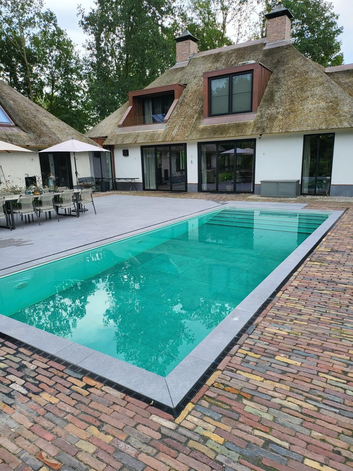 Rechthoekig turquoise zwembad voor een huis met een rieten dak, omgeven door een bakstenen terras. MagicPool