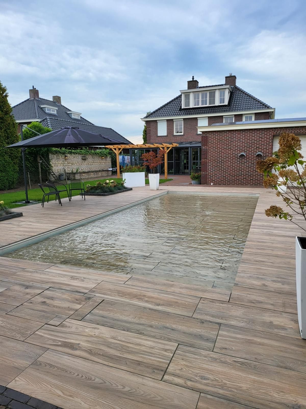 Rechthoekig zwembad (MagicPool) met houten terras, omgeven door een huis en weelderig groen onder een bewolkte hemel.
