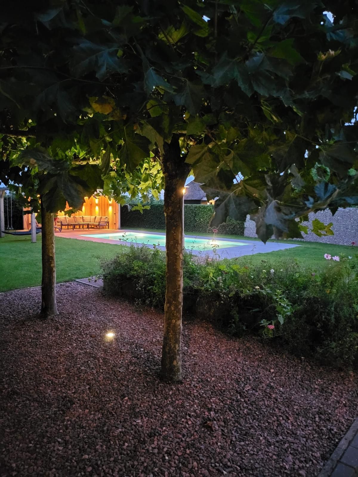 Bomen met verlichte kruinen, die uitkijken op een gebouw en een gazon met bloembedden en een verlicht pad en een MagicPool.