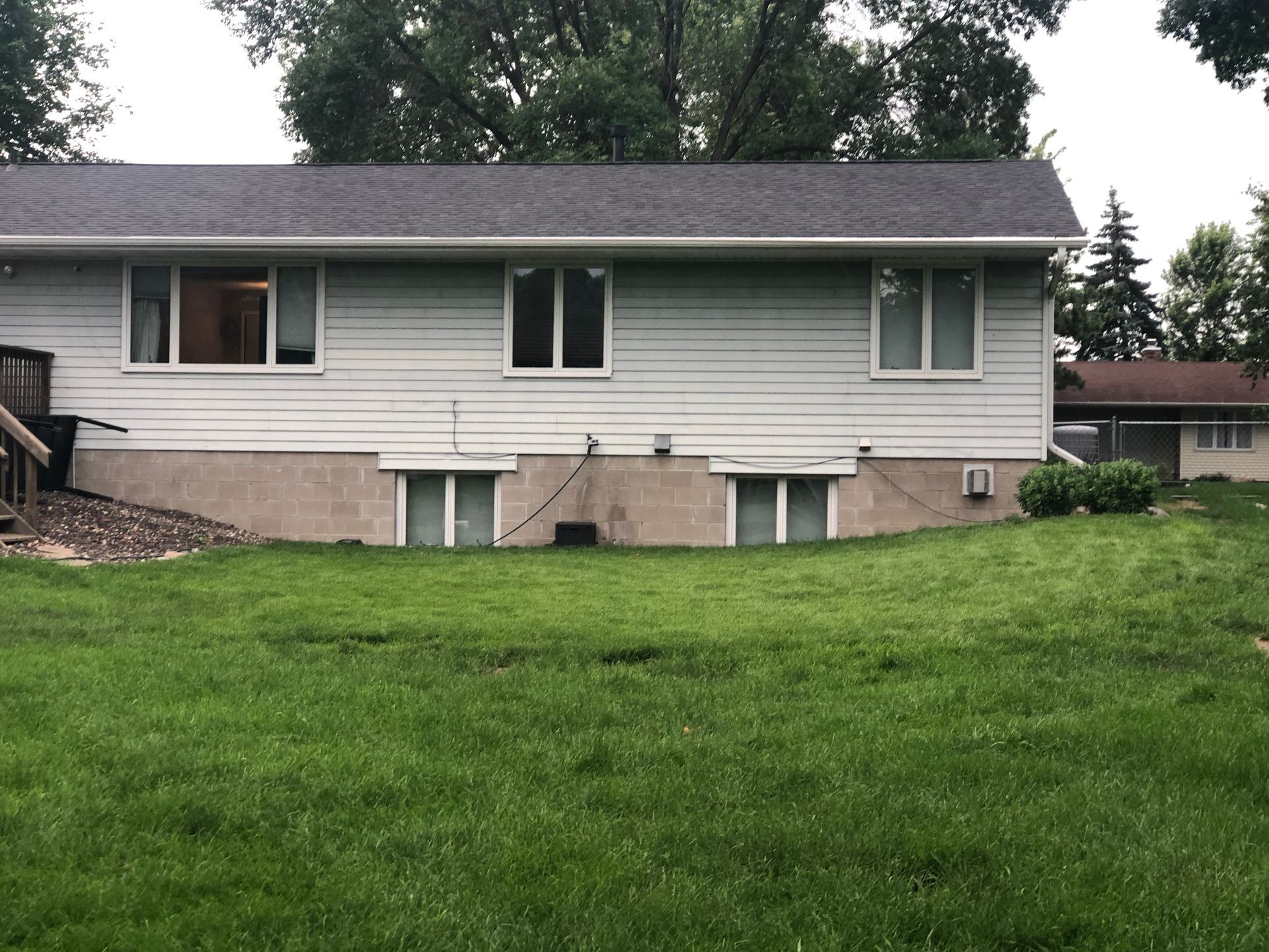 Suburban house with white siding, basement windows, and a large green lawn under overcast skies