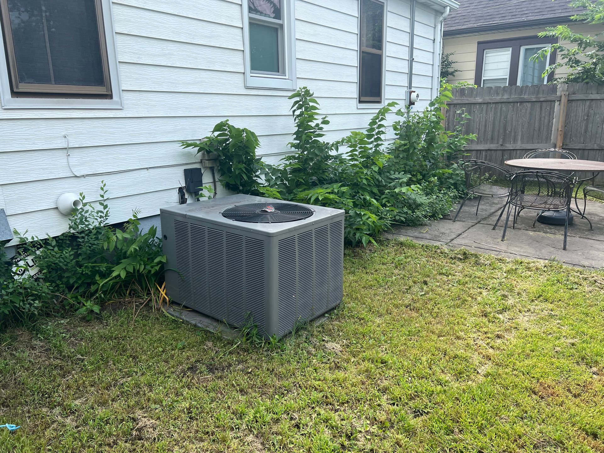 Outdoor HVAC unit beside a white house, with a grassy yard and patio furniture nearby