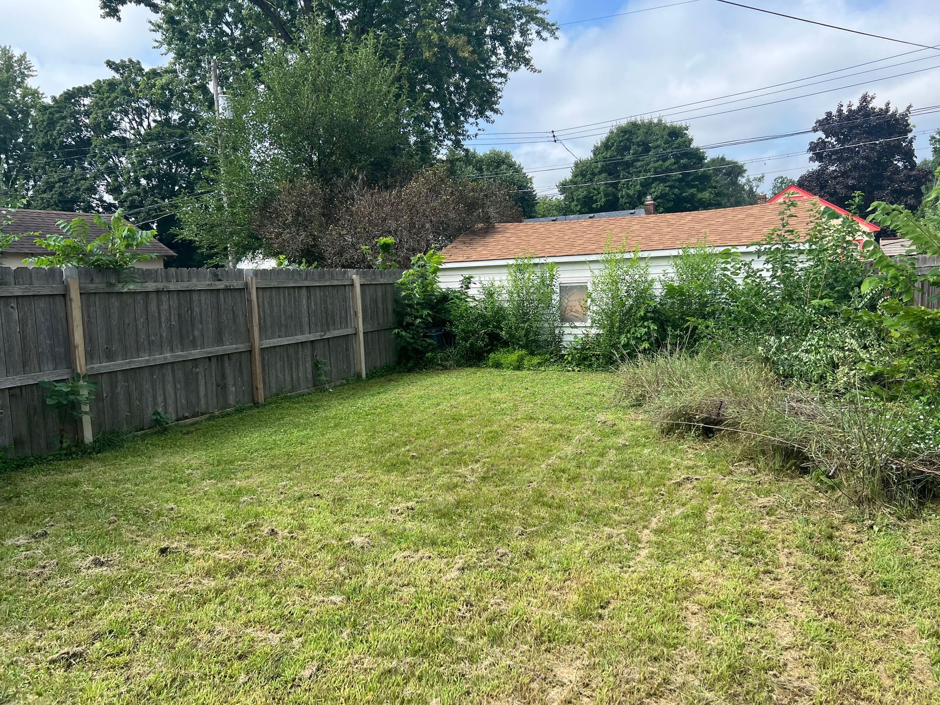 Backyard with a grassy lawn, wooden fence, shrubs, and a small white outbuilding under a cloudy sky