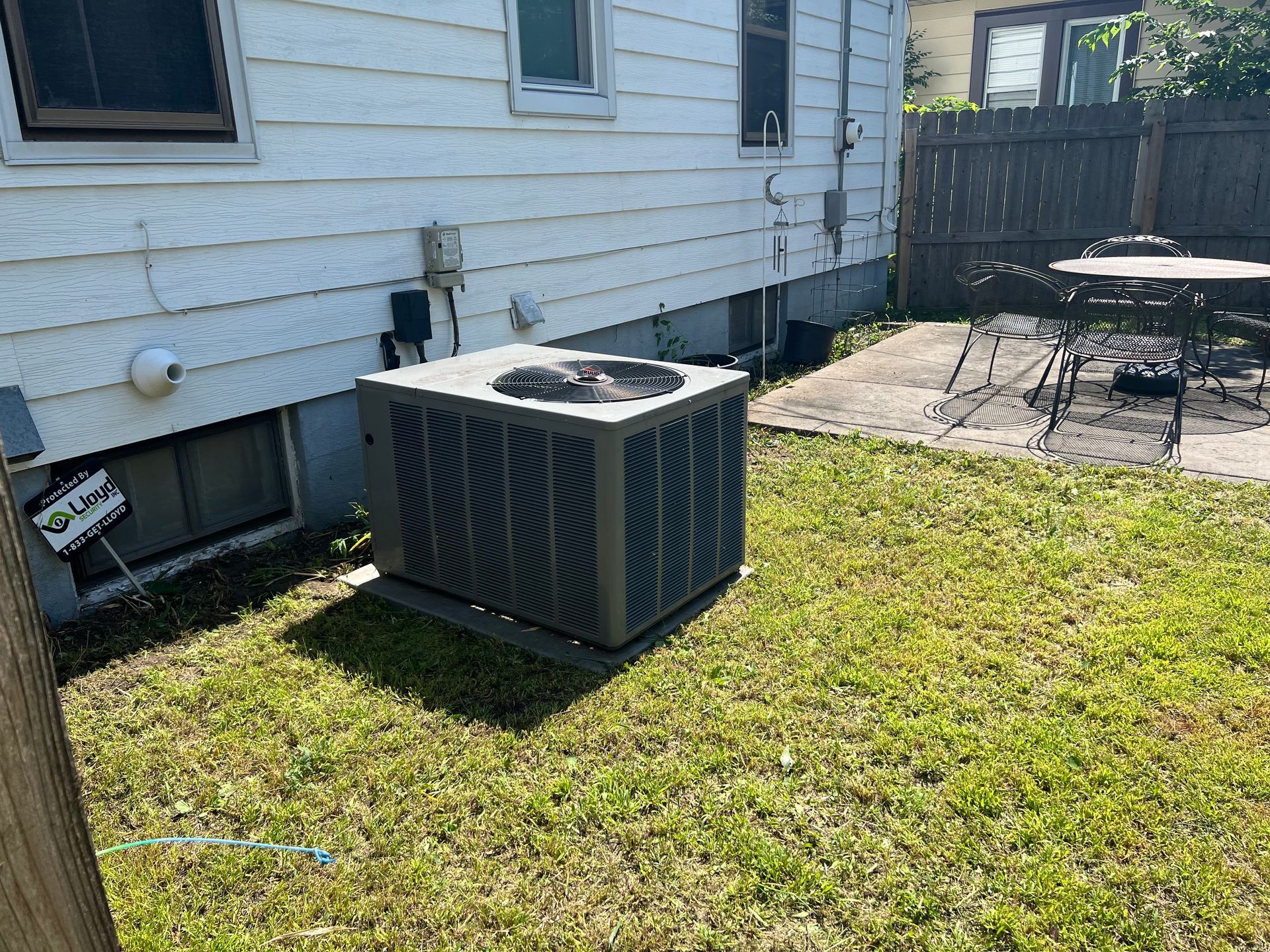 Outdoor HVAC unit beside a white house, on a grassy yard near a patio and wooden fence.