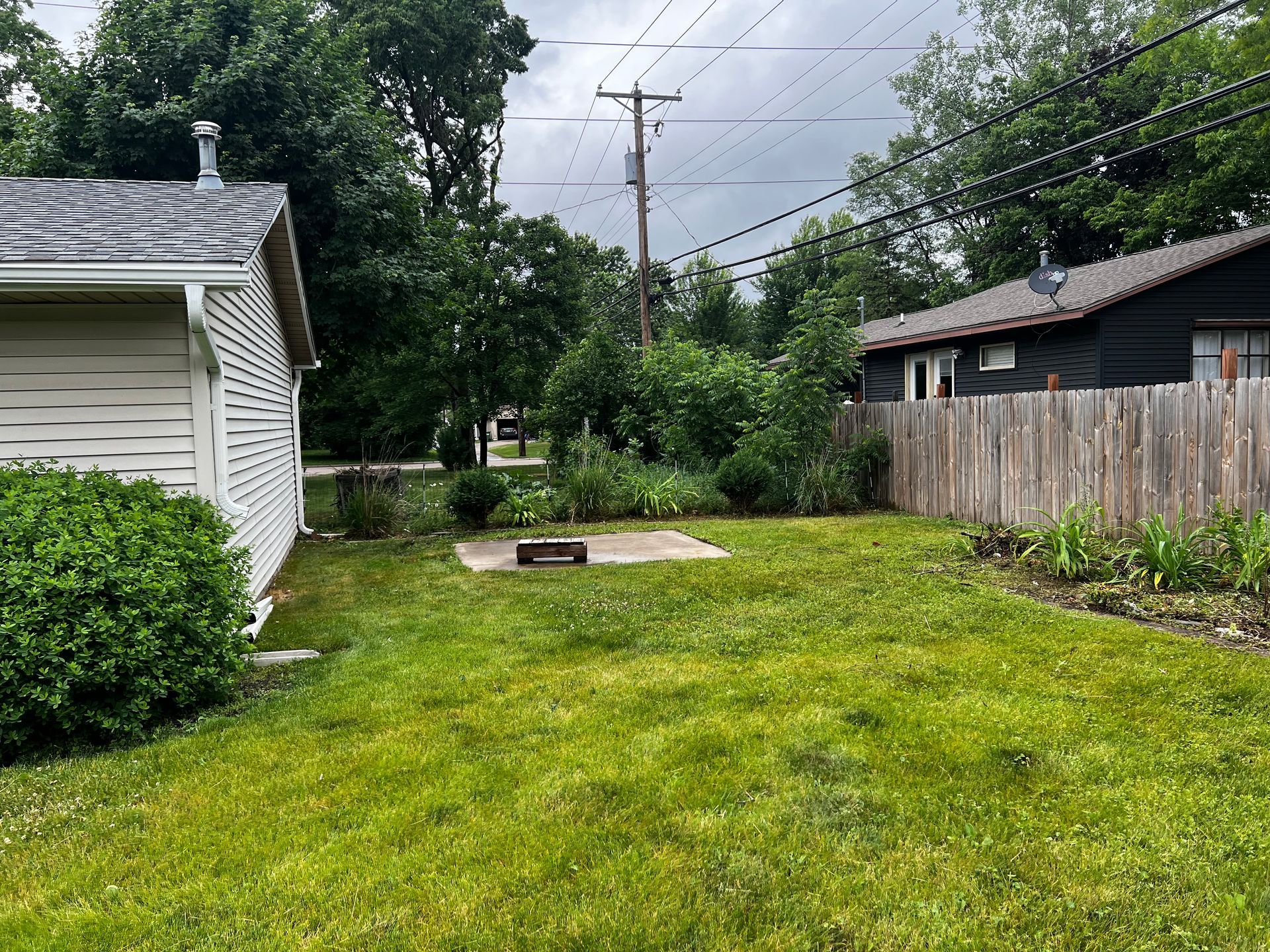 Narrow grassy backyard between houses with a fence, utility pole, and cloudy sky