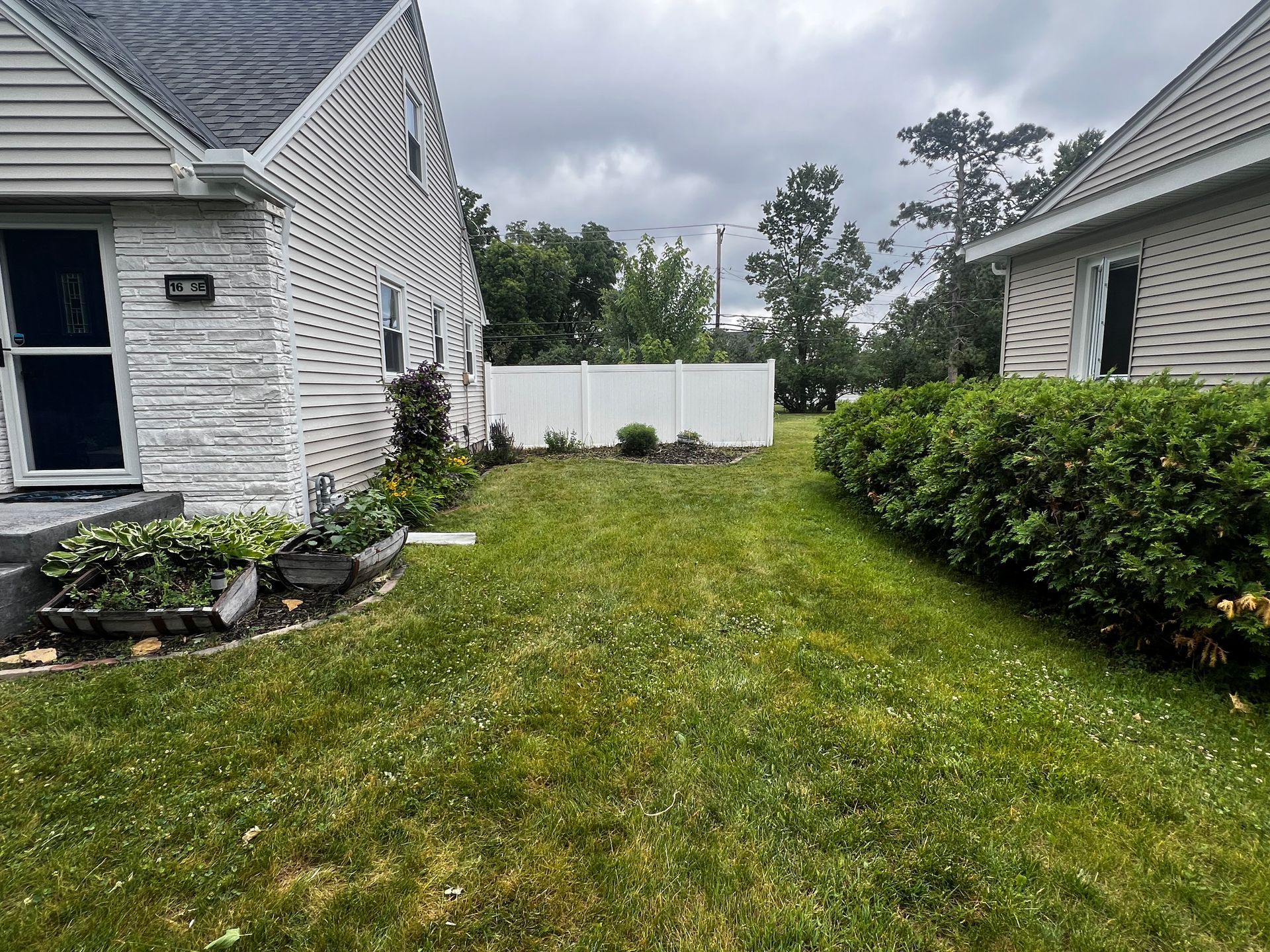 Narrow grassy side yard between two white houses, with shrubs and a white fence under cloudy skies