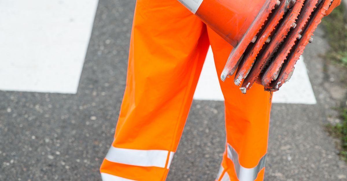 Worker in bright orange pants carrying stacked orange traffic cones on a road crossing