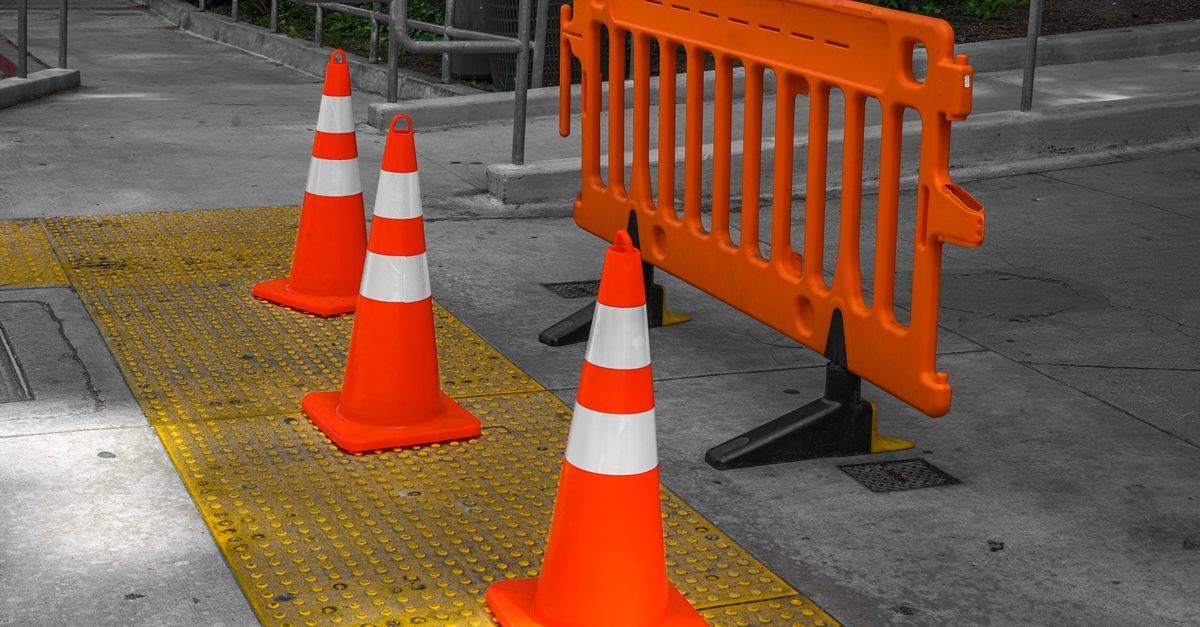 Orange traffic cones and barrier blocking a sidewalk section with yellow tactile paving, beside a wet road.