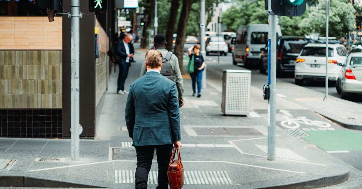 Person waiting to cross a city street at a crosswalk, holding a bag, with cars and pedestrians nearby