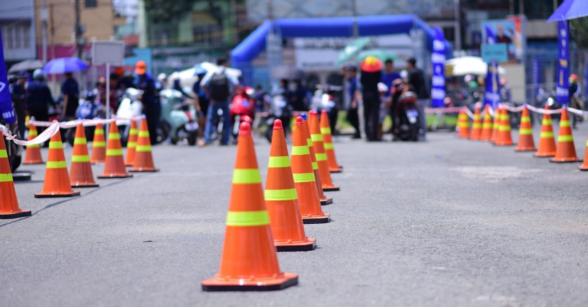 Orange traffic cones line a street, with blurred pedestrians and tents in the background.