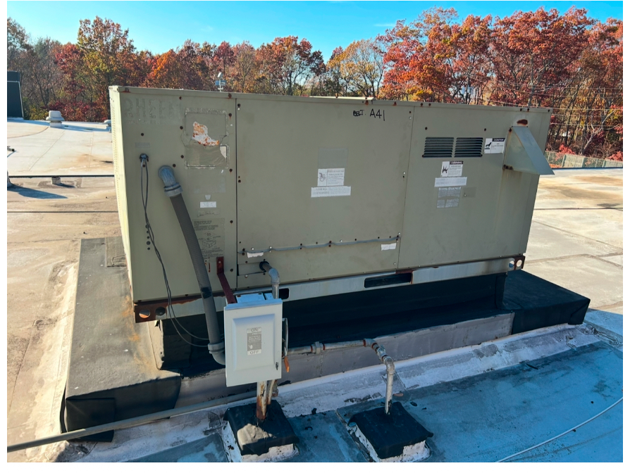 A large, beige commercial HVAC unit sits on a flat rooftop against a backdrop of trees with autumn foliage.