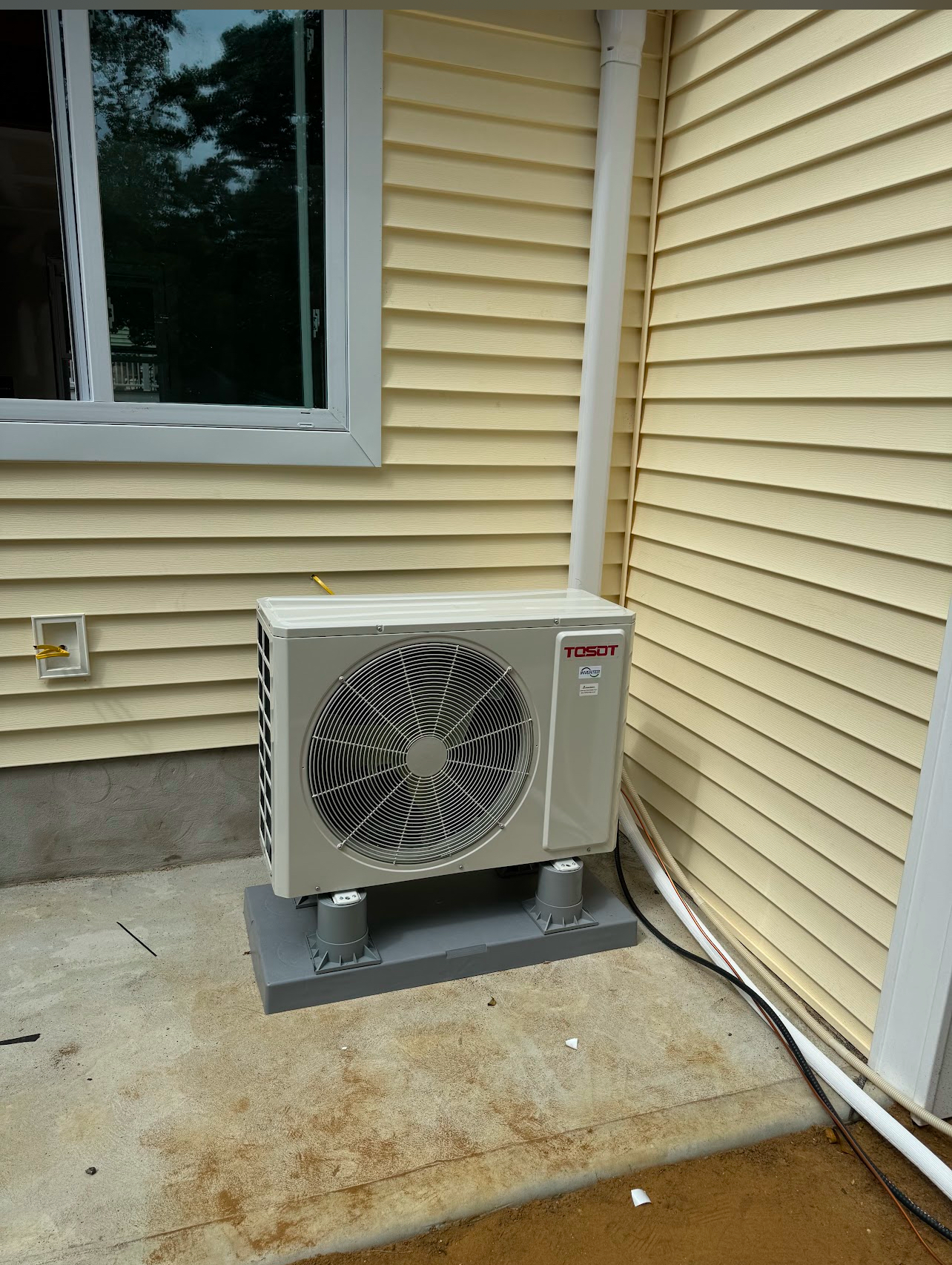 A beige outdoor heat pump unit sits on a grey plastic stand against a yellow-sided house near a window.