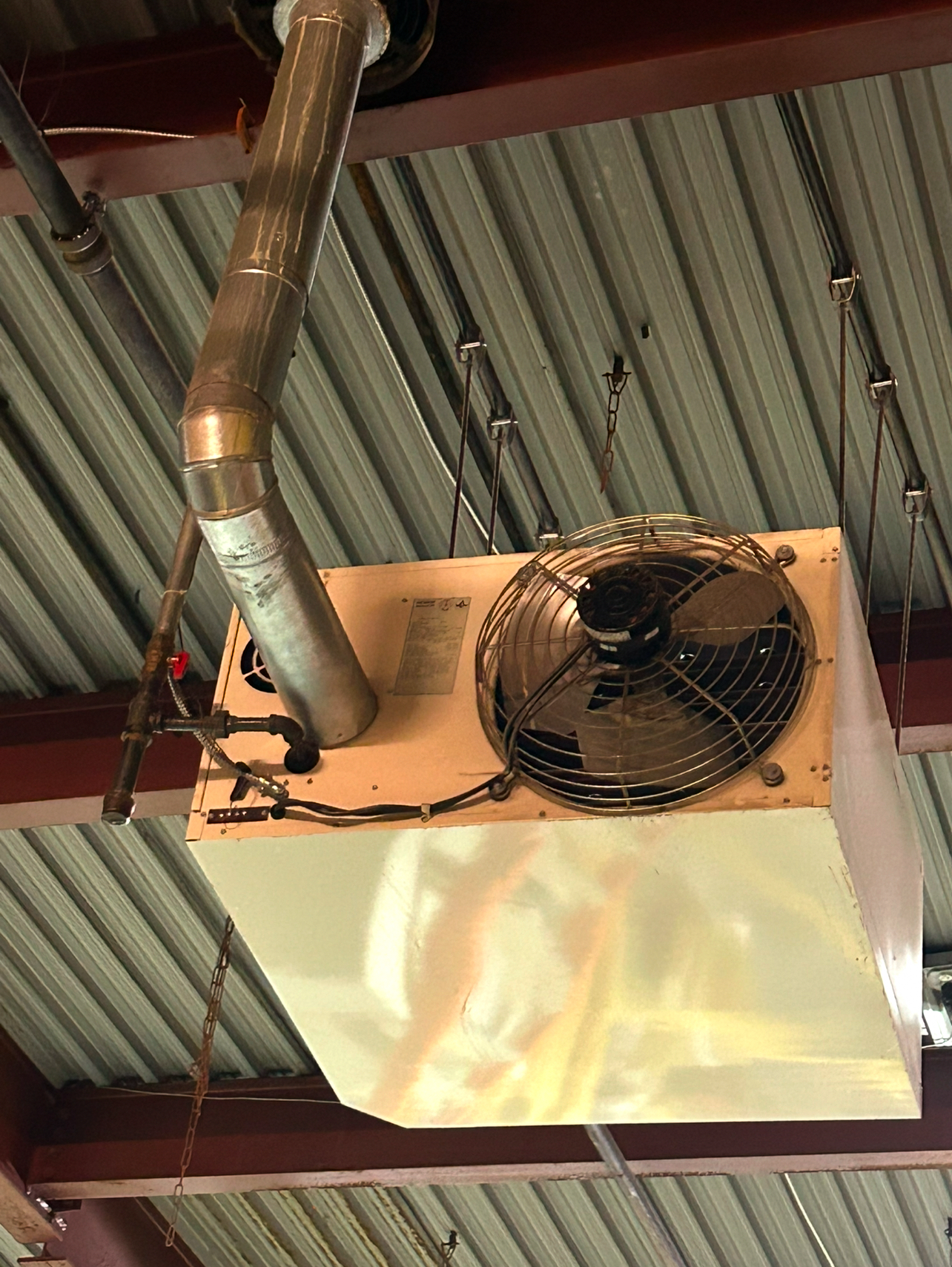 A beige industrial unit heater with a black fan mounted to a ceiling with a metal exhaust pipe.