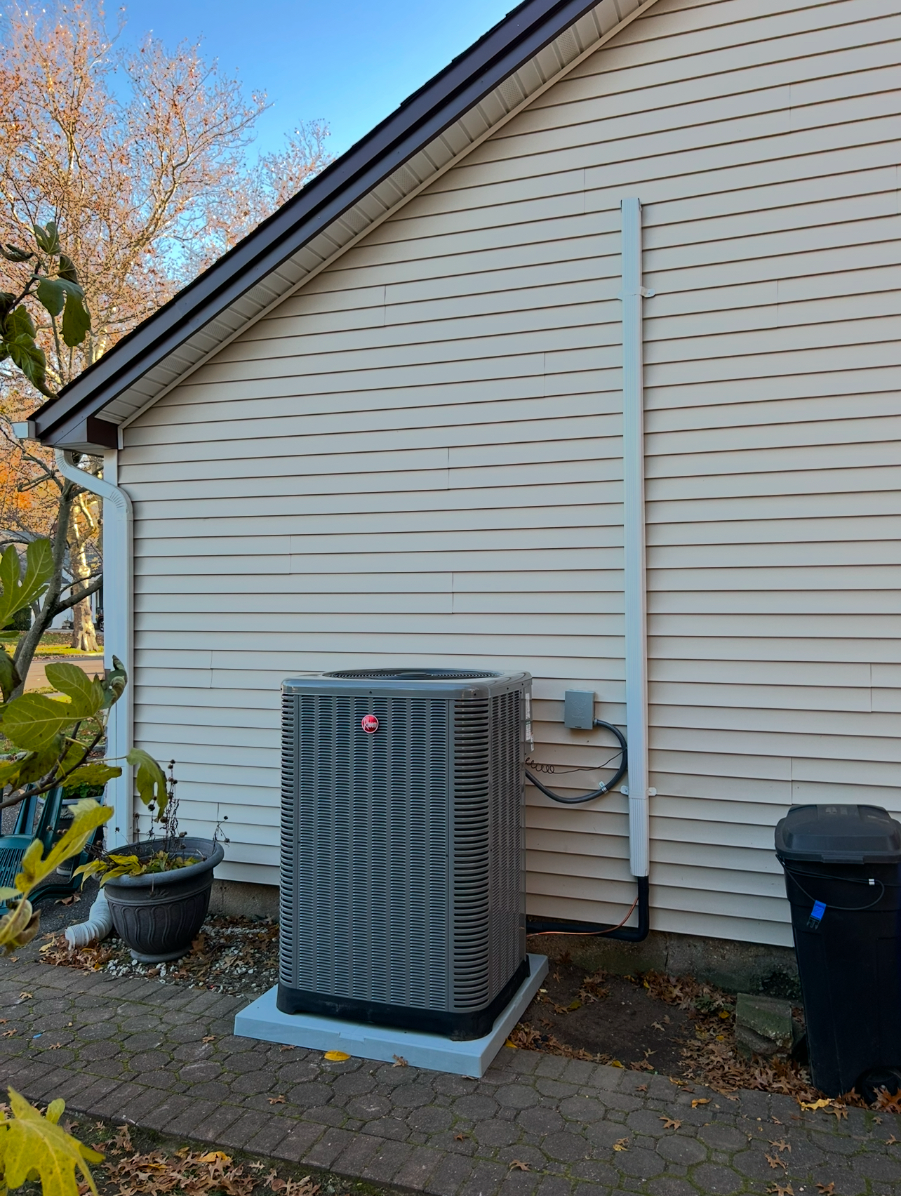 A grey HVAC unit sits on a concrete pad against the light-colored vinyl siding of a house exterior.