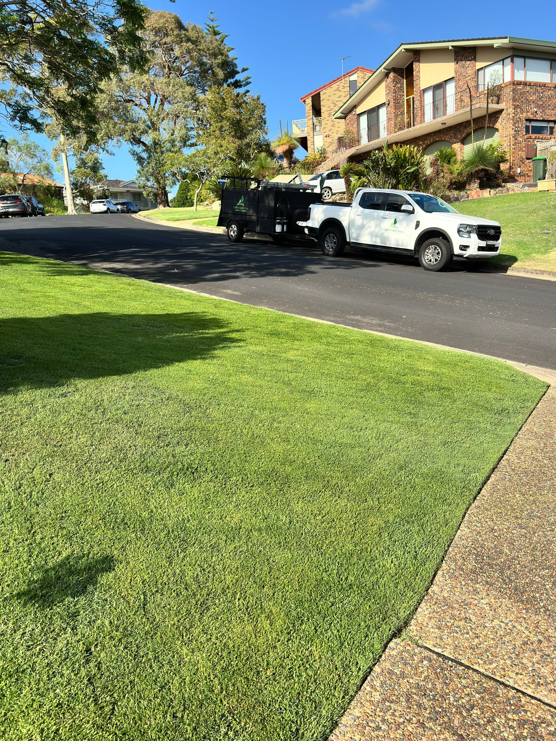 Sunny suburban street with parked pickup trucks, brick townhouses, green lawn, and sidewalk