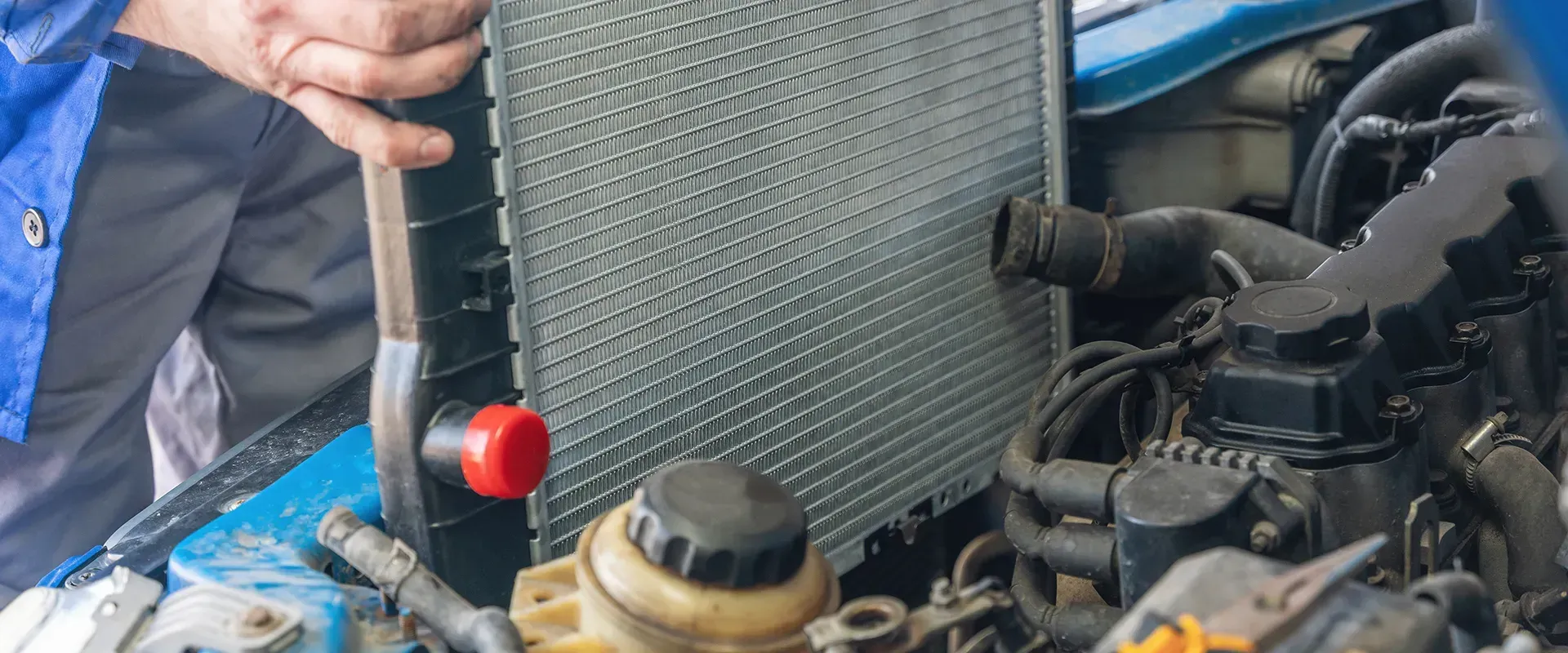 A mechanic works on a car engine, holding a radiator. The setting is a car repair shop.