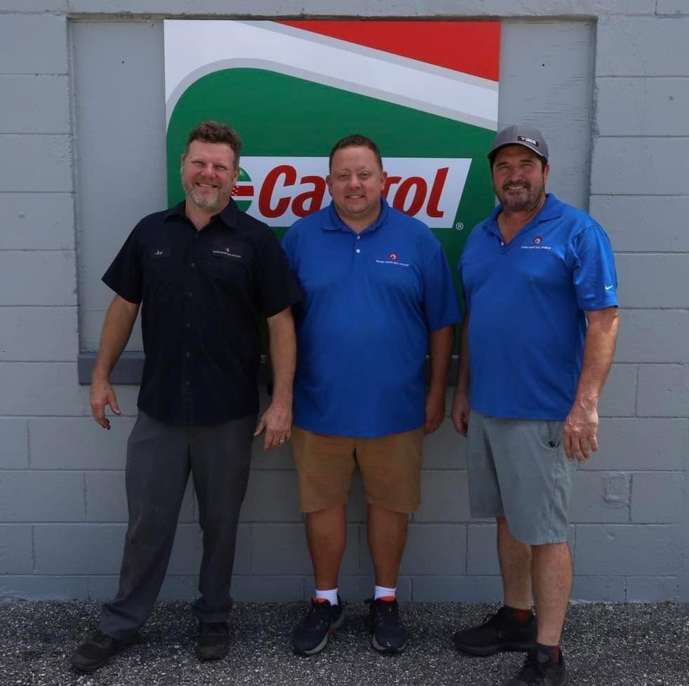 Three men stand in front of a Castrol sign on a gray brick wall. Two in blue shirts, one in black.