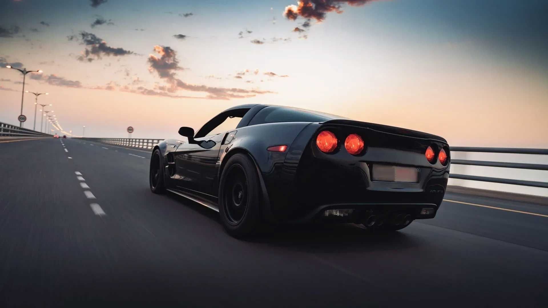 Black sports car driving on a bridge at sunset with orange and blue sky.