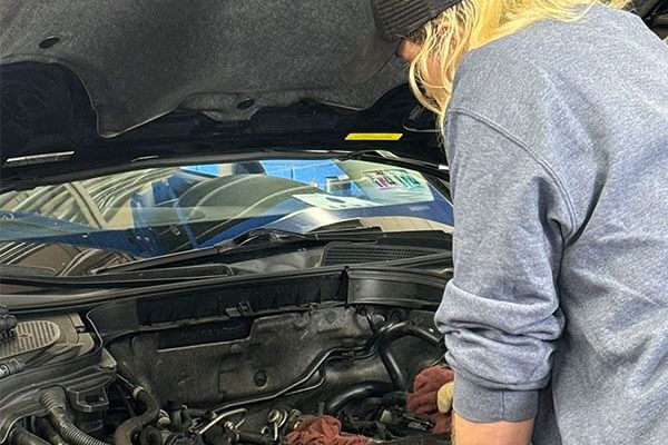 Person with blonde hair looks at car engine. Engine bay is open.