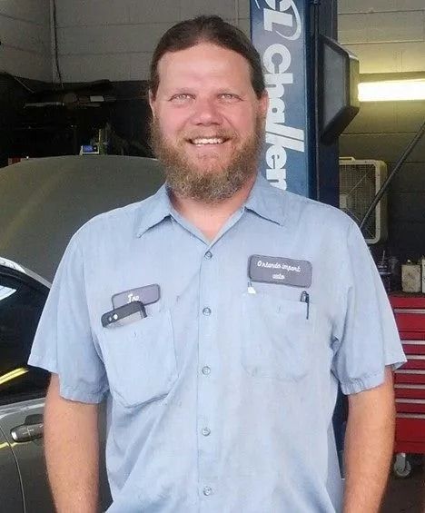 Mechanic in a blue shirt smiling, standing in a garage, next to a car.