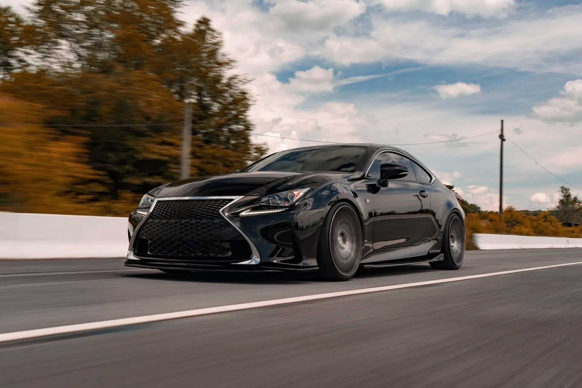 Black Lexus RC coupe driving on a highway, blurred background of trees and sky.