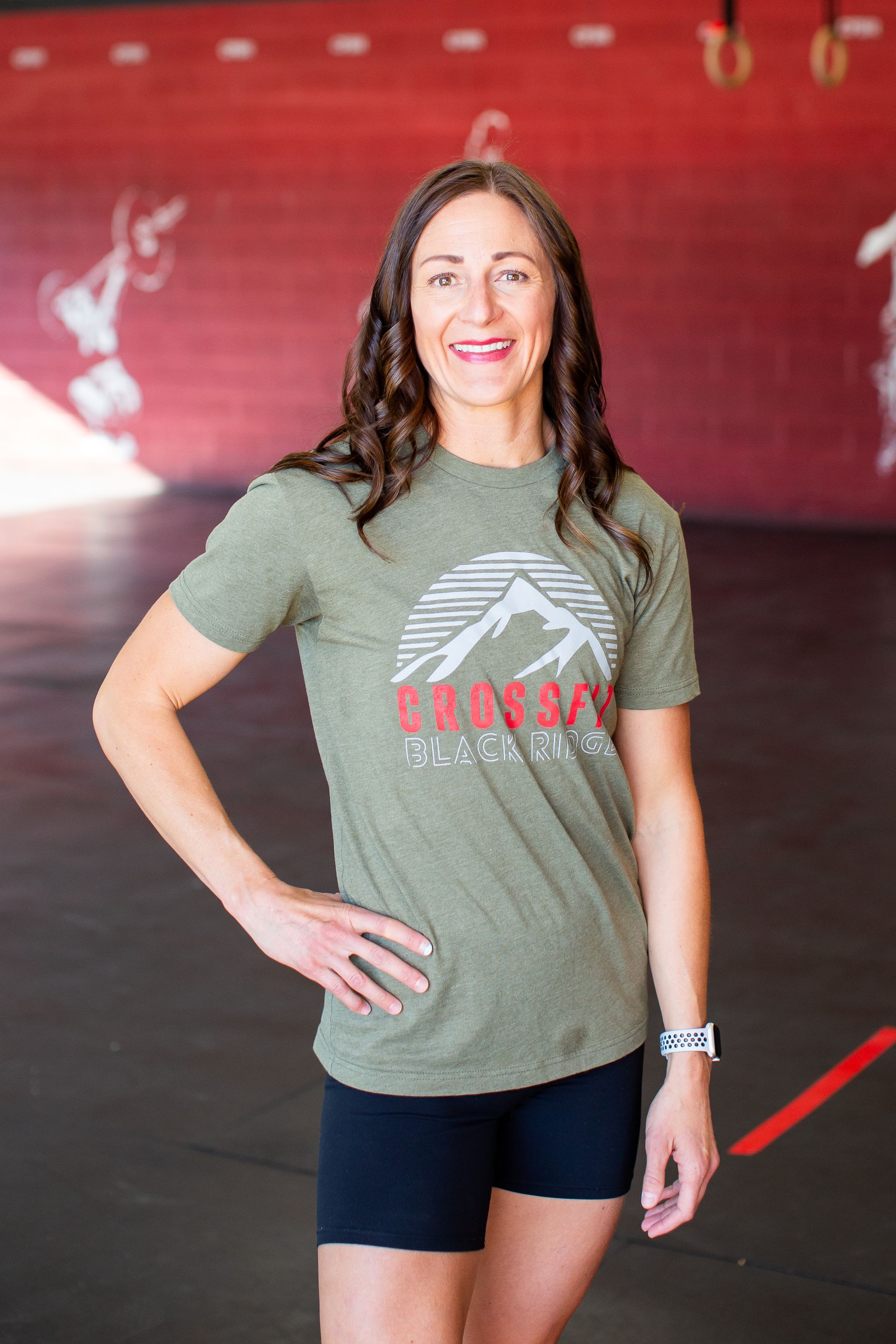 A woman wearing a green t-shirt and black shorts is standing in a gym with her hands on her hips.