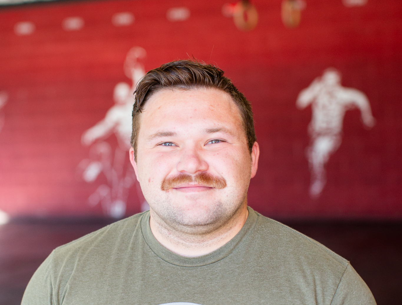 A man with a mustache is smiling in front of a red wall.