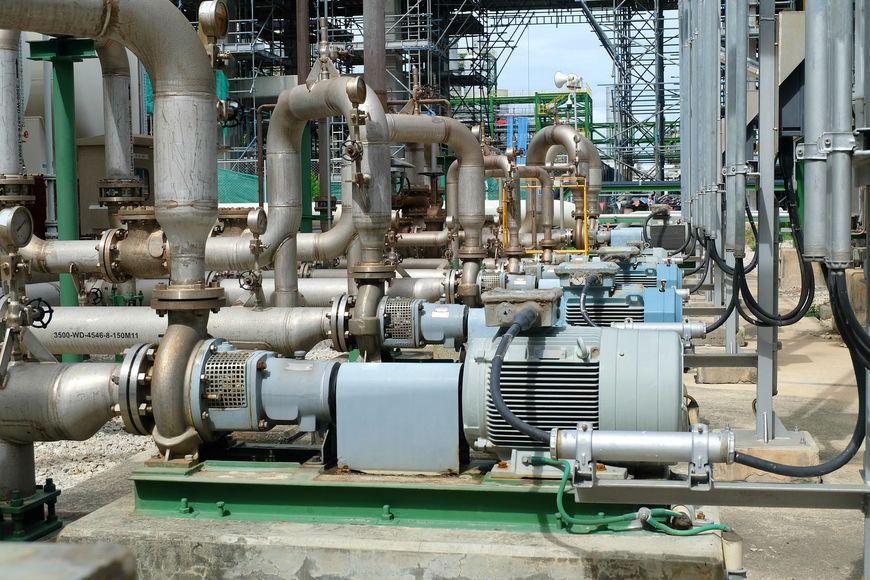 A row of industrial centrifugal pumps and electric motors connected by metal piping in an outdoor refinery setting.