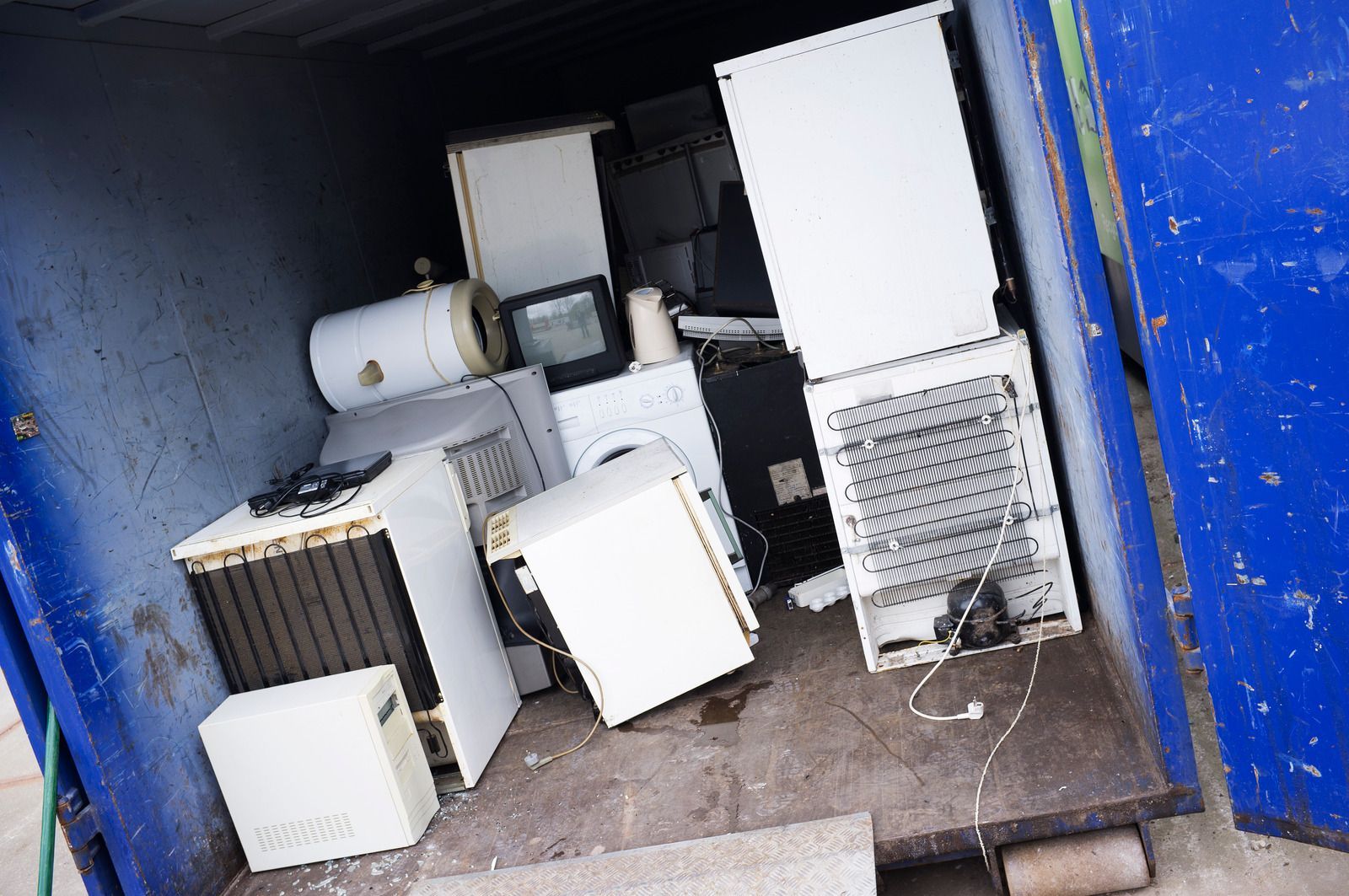 Blue dumpster filled with discarded appliances: refrigerator, water heater, and TV.