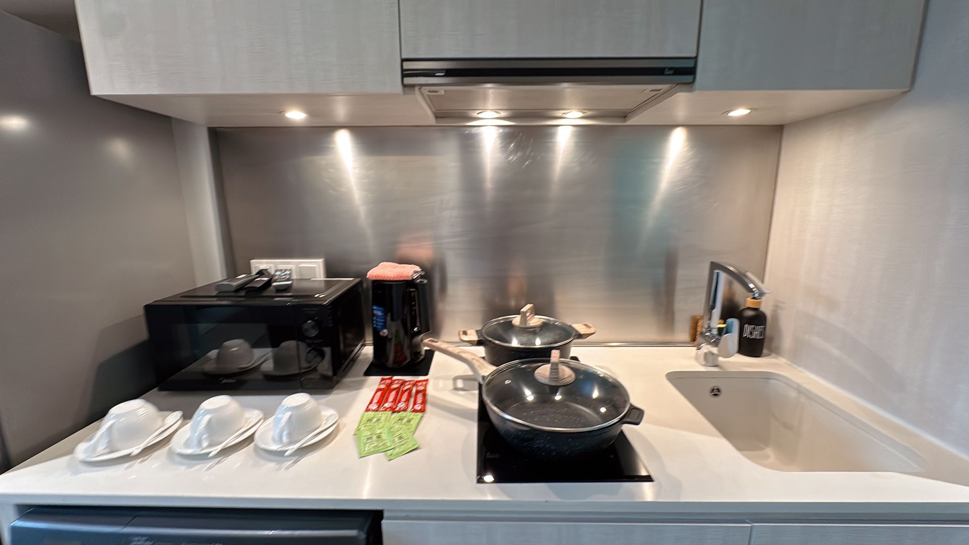 A compact kitchen with a microwave, coffee maker, cooktop, and sink. Stainless steel backsplash.