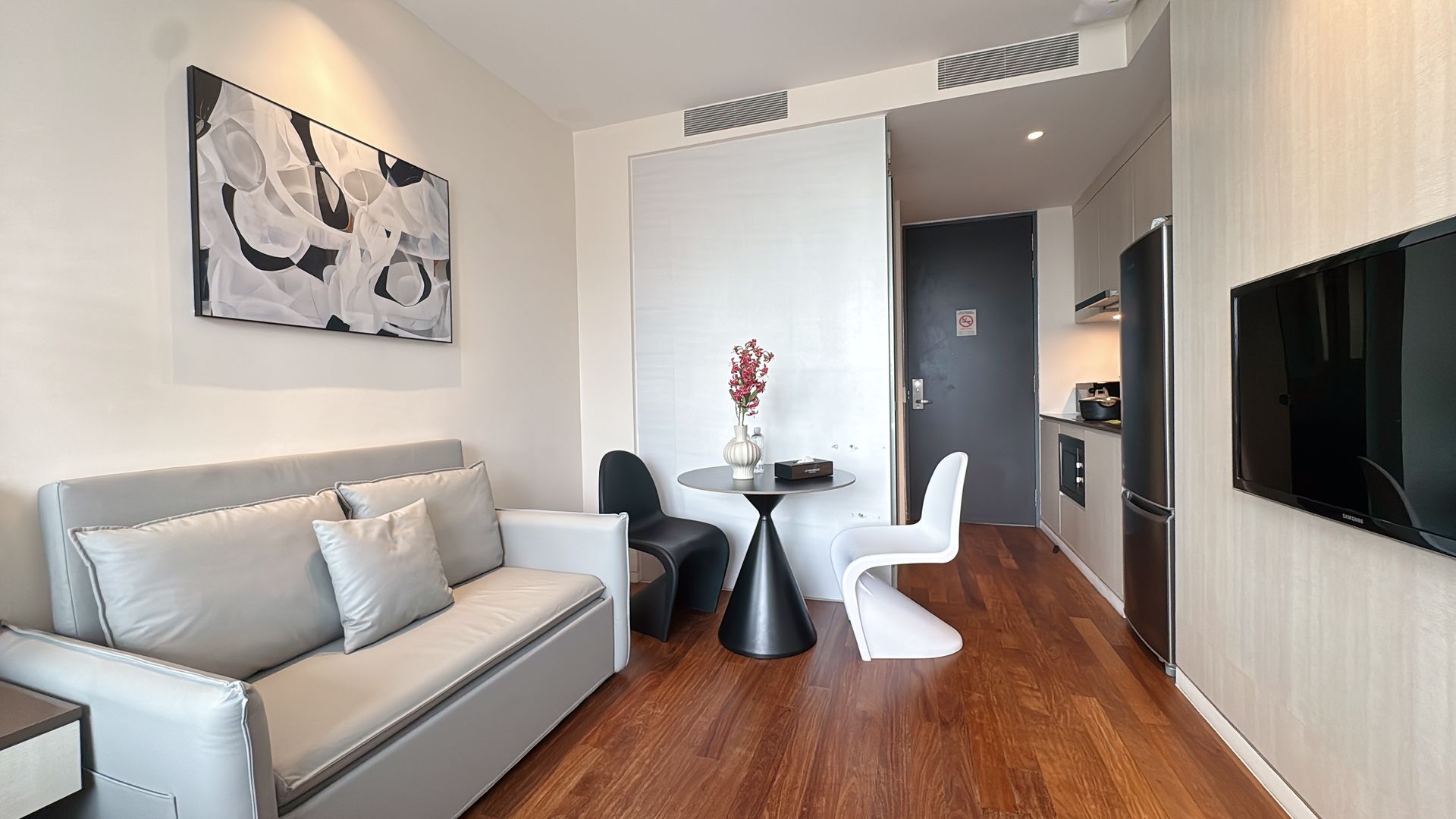 Modern studio apartment with sofa, table, kitchen, and TV; wooden floor and artwork.