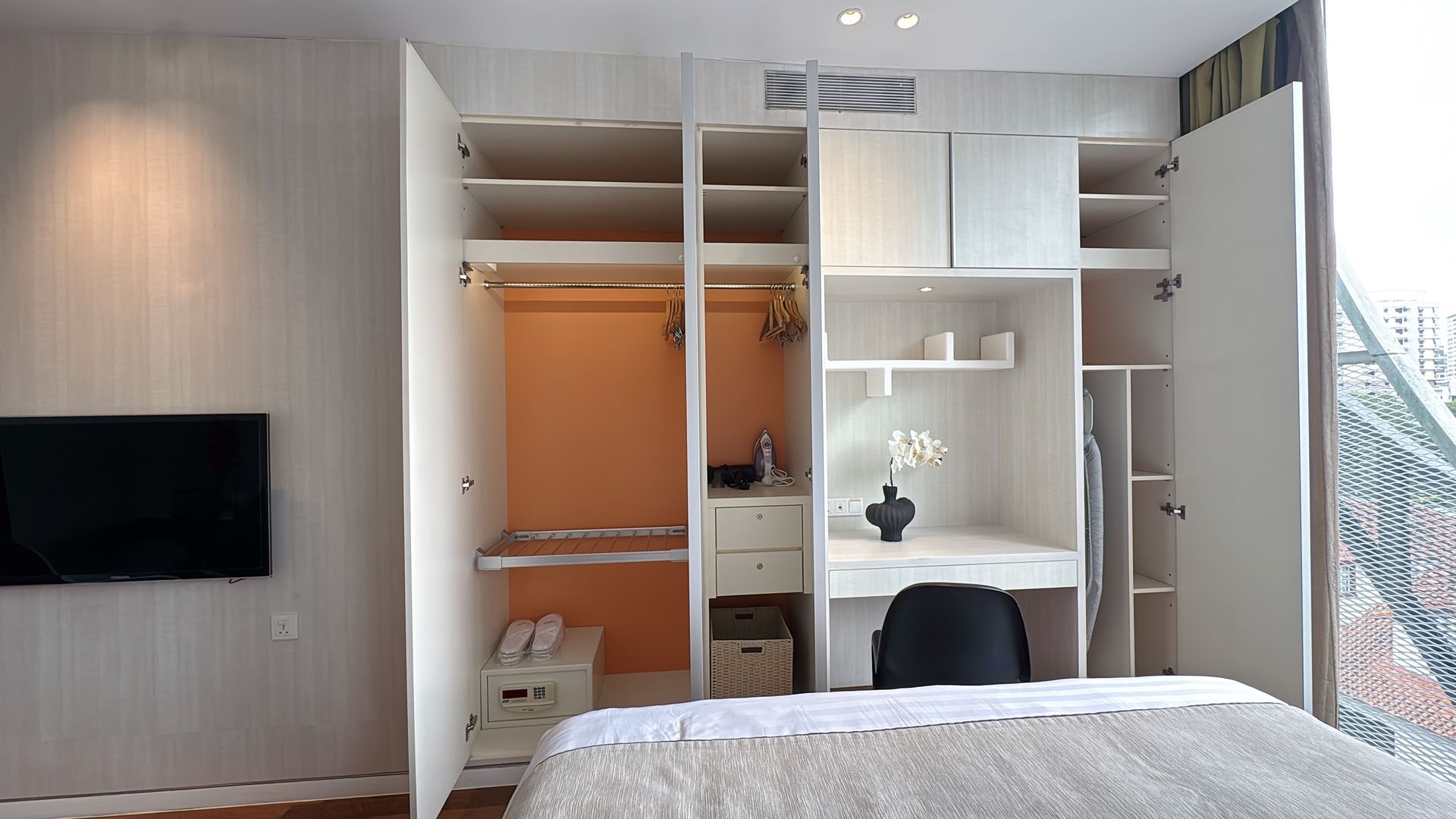 A white open closet with a desk, shelves, and clothing rods. Bedroom with a TV on the wall.