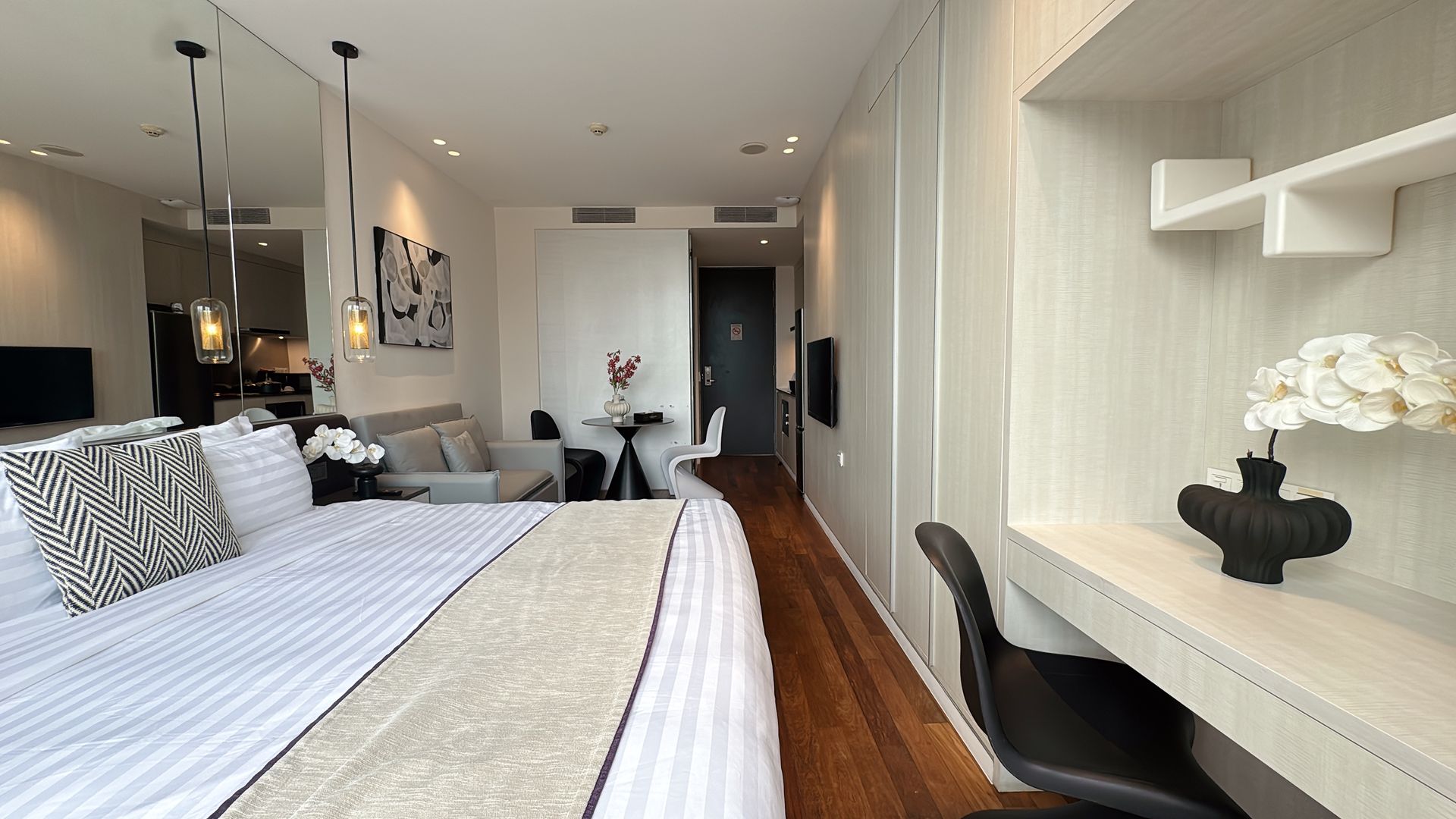 Modern studio apartment with a bed, desk, and seating area. White and neutral tones.