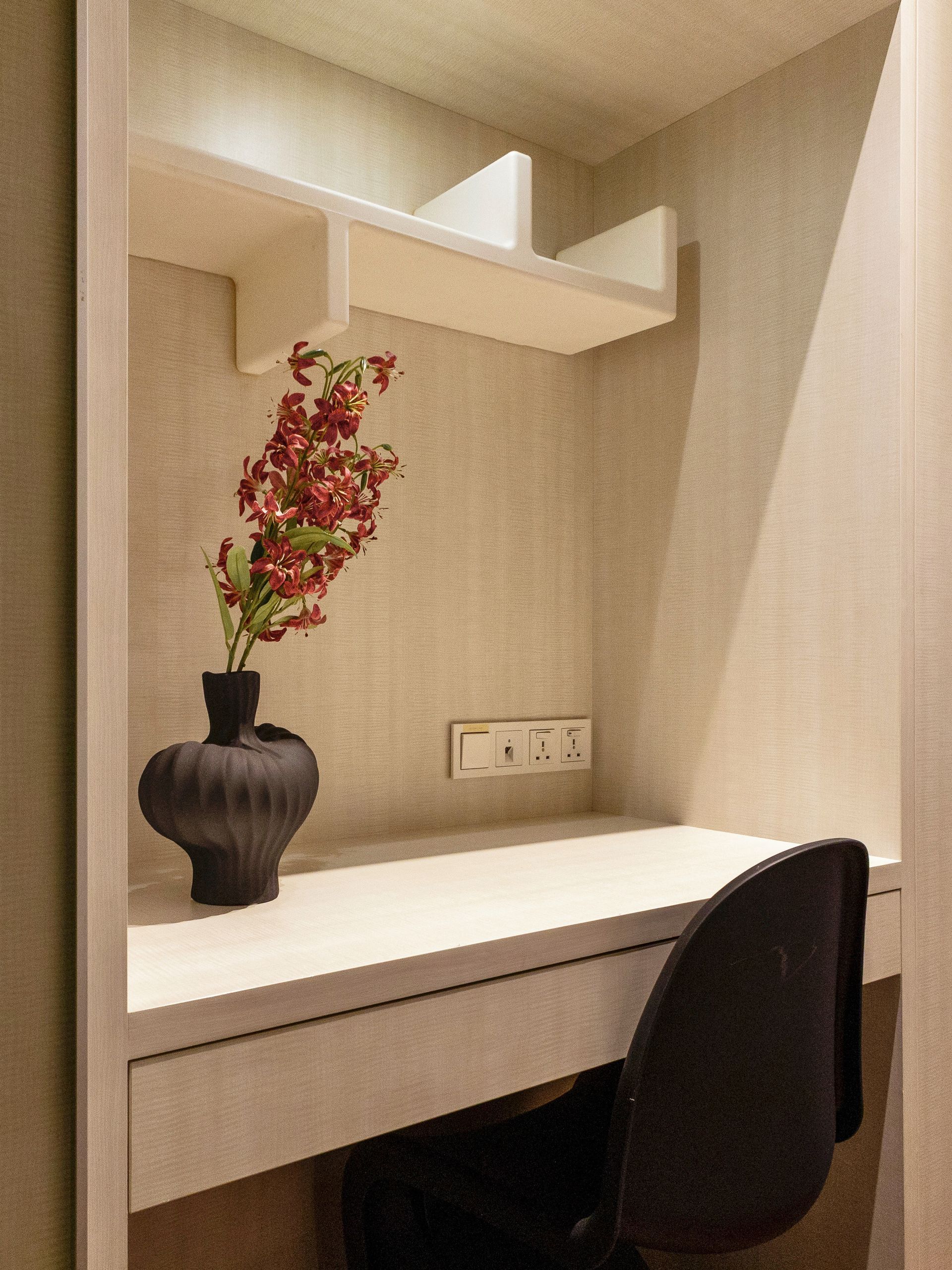 Built-in desk with black chair, vase of red flowers. Beige shelves, textured wall, and outlets.