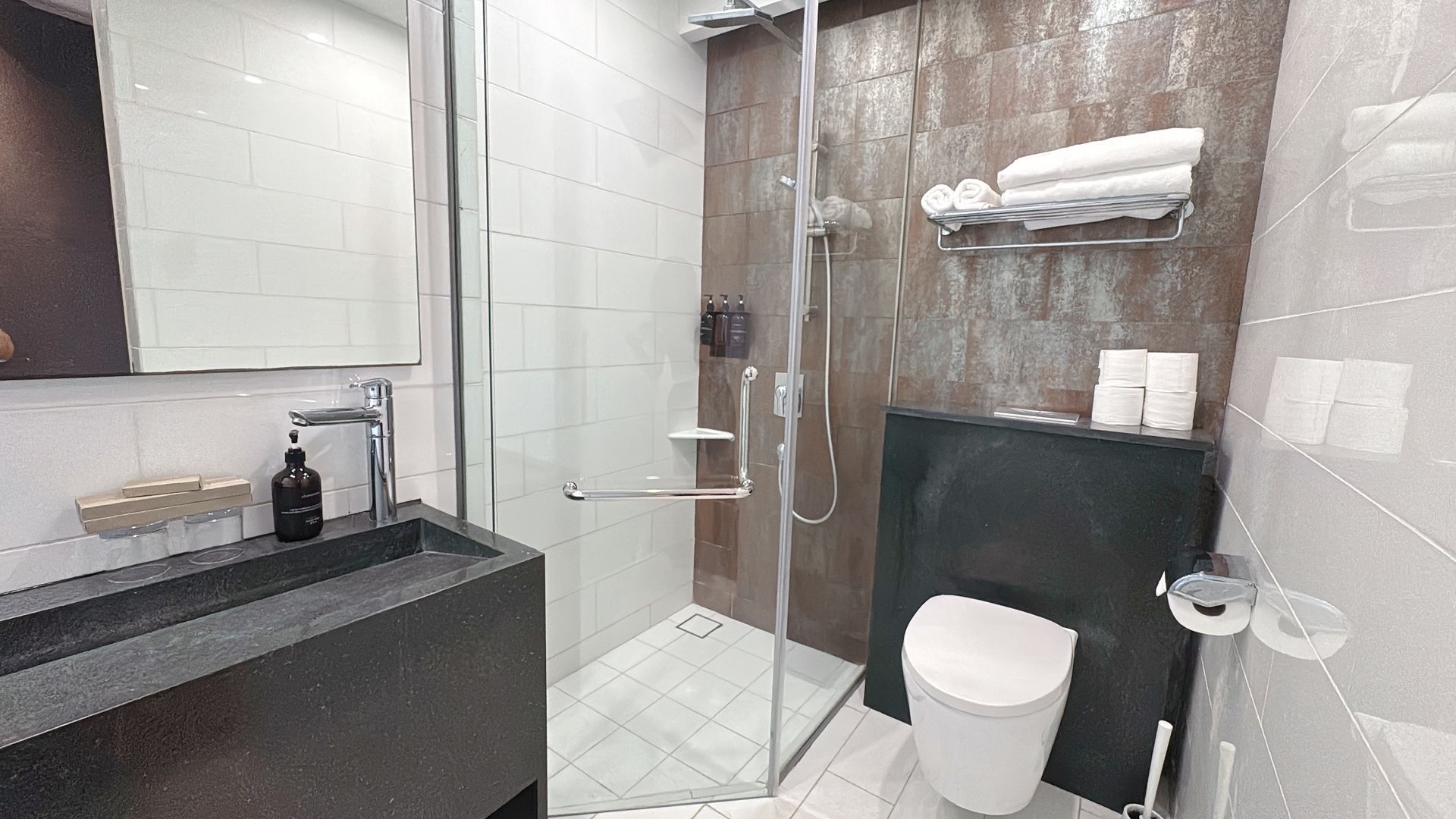 Modern bathroom with dark sink, glass shower, toilet, and brown tiled wall.