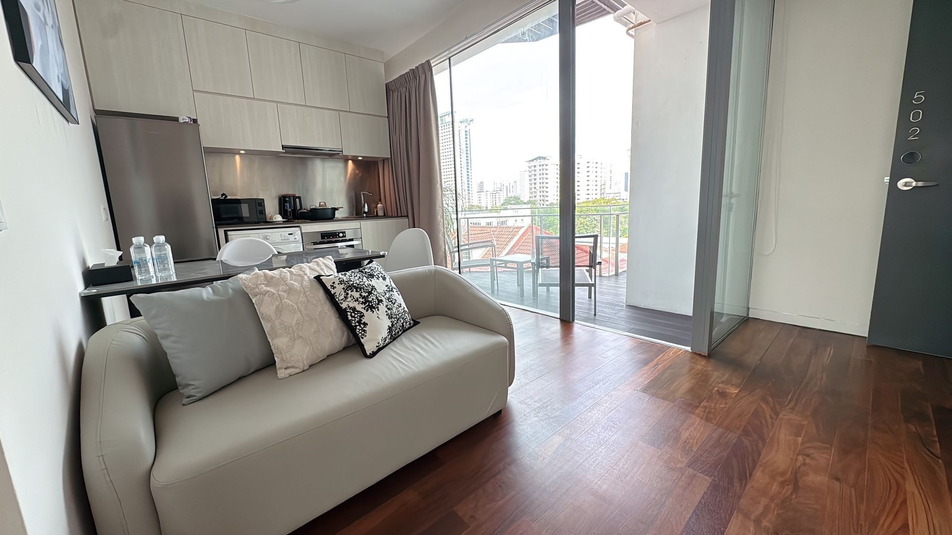 A small, modern apartment with a couch, open balcony, and a small kitchenette.