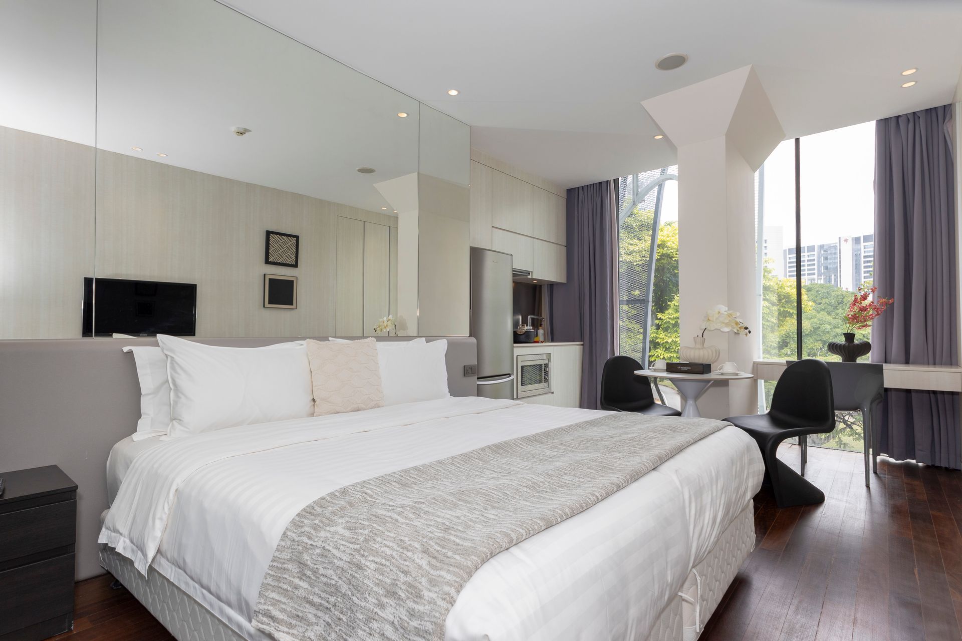 Modern bedroom with large bed, full mirror, and floor-to-ceiling window overlooking a city.