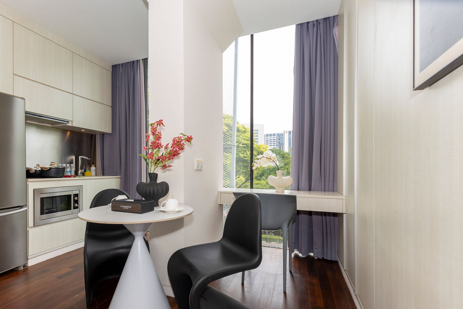 Small, modern kitchen area with table, black chairs, large window with purple curtains.