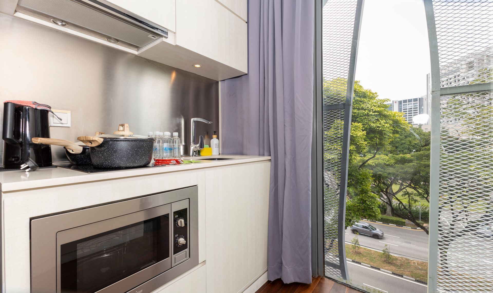 Kitchen with microwave, cooking pot, and window overlooking a road with trees and a car.