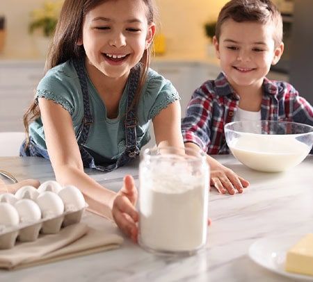 Two kids baking in a kitchen with flour, eggs, milk, and butter.