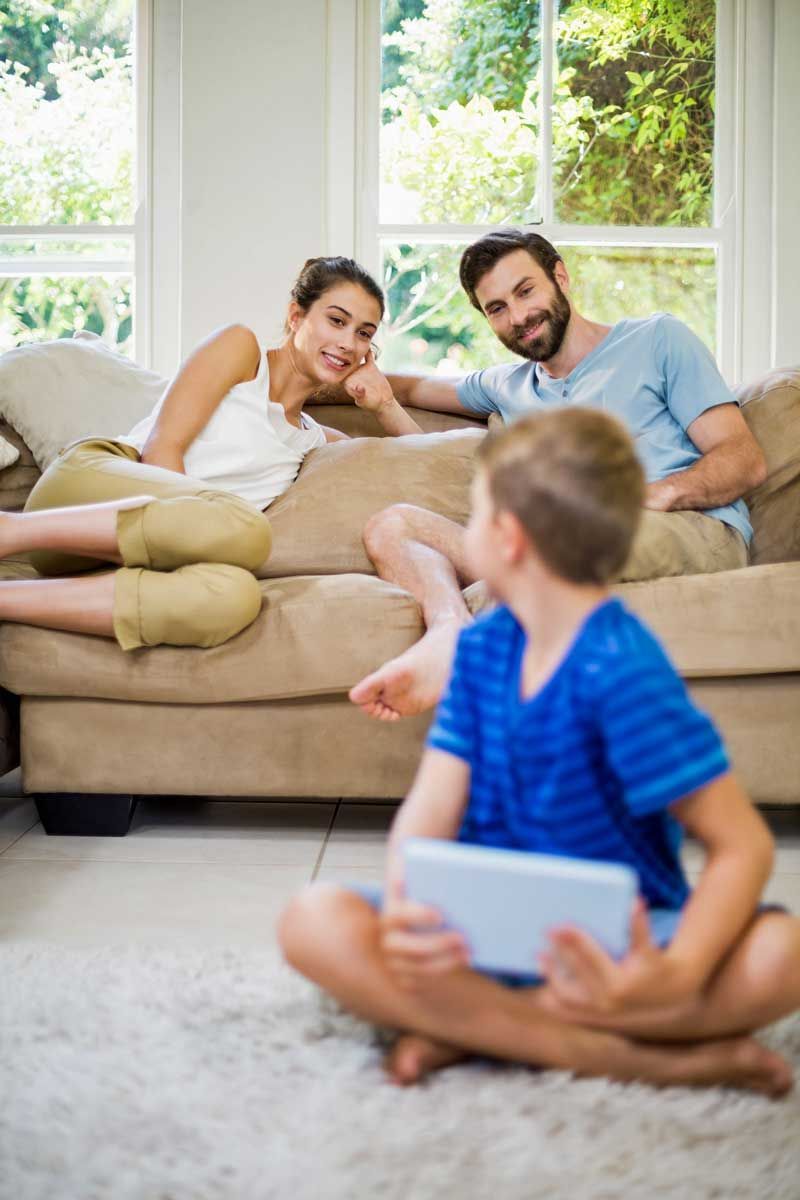 A young boy sits on a rug, using a tablet, while his parents relax on a couch in a sunny living room.