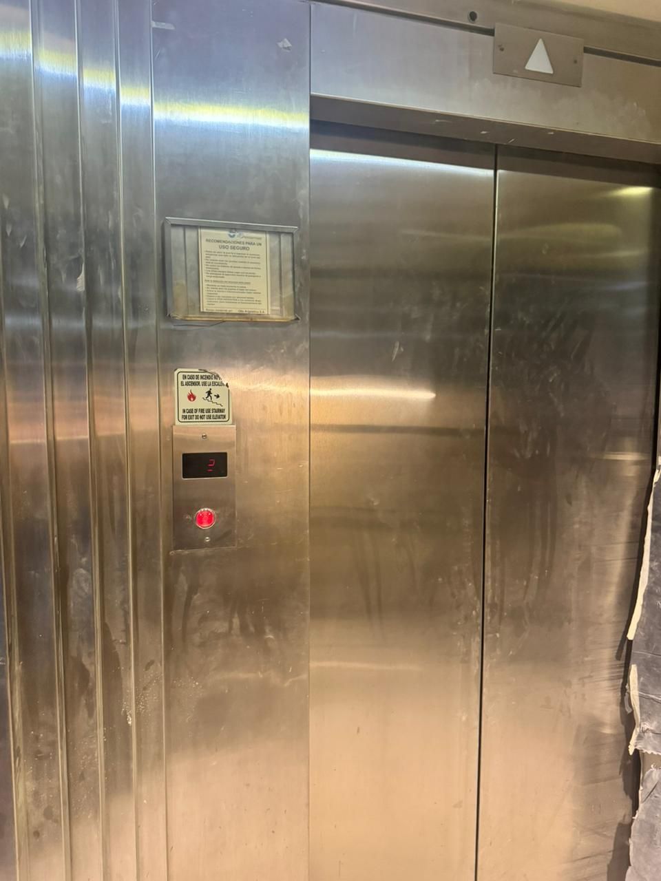 Puertas de ascensor cerradas de acero inoxidable con panel de botones de llamada y aviso publicado.