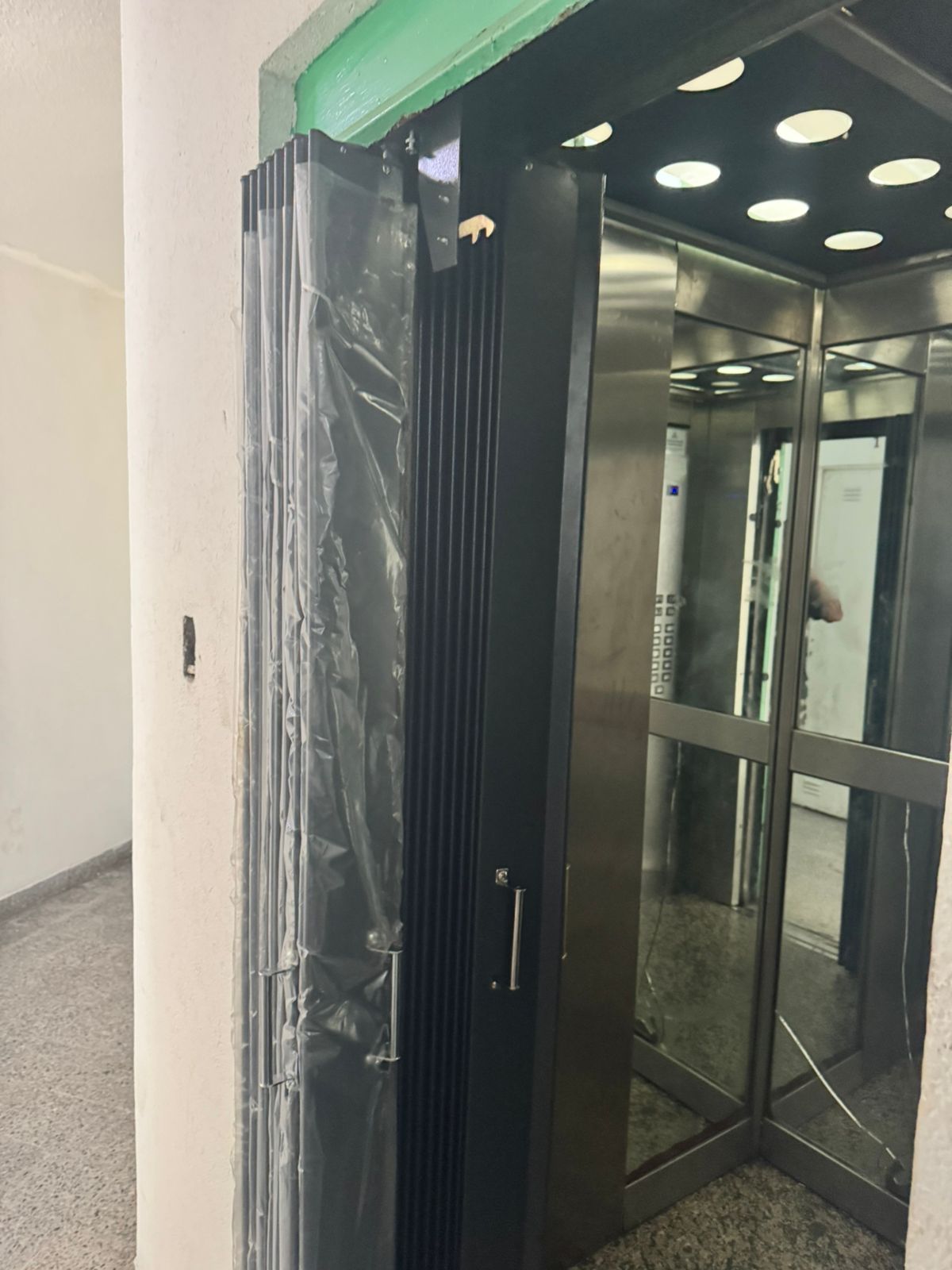 Ascensor con puertas de cristal y marco negro en un pasillo, parcialmente cubierto con una lámina de plástico.