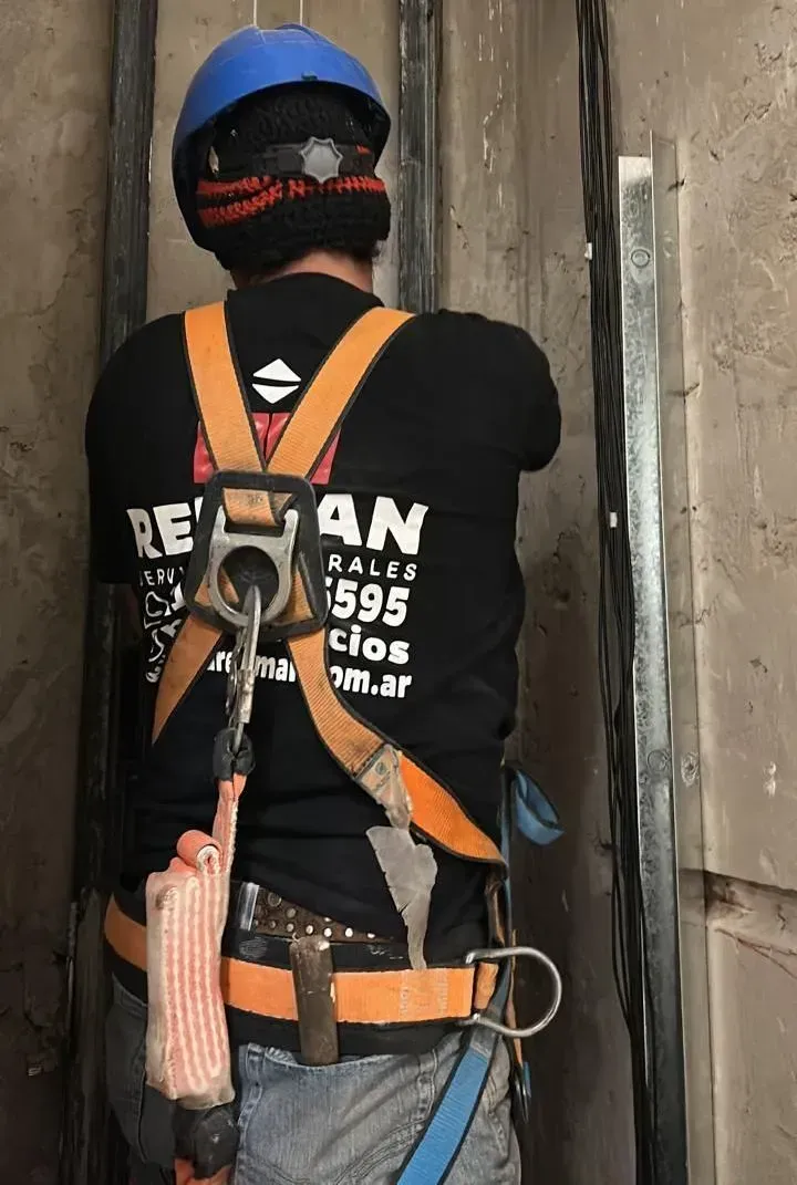 Un trabajador con casco azul y arnés naranja desciende en rápel por una pared de hormigón dentro de un edificio.