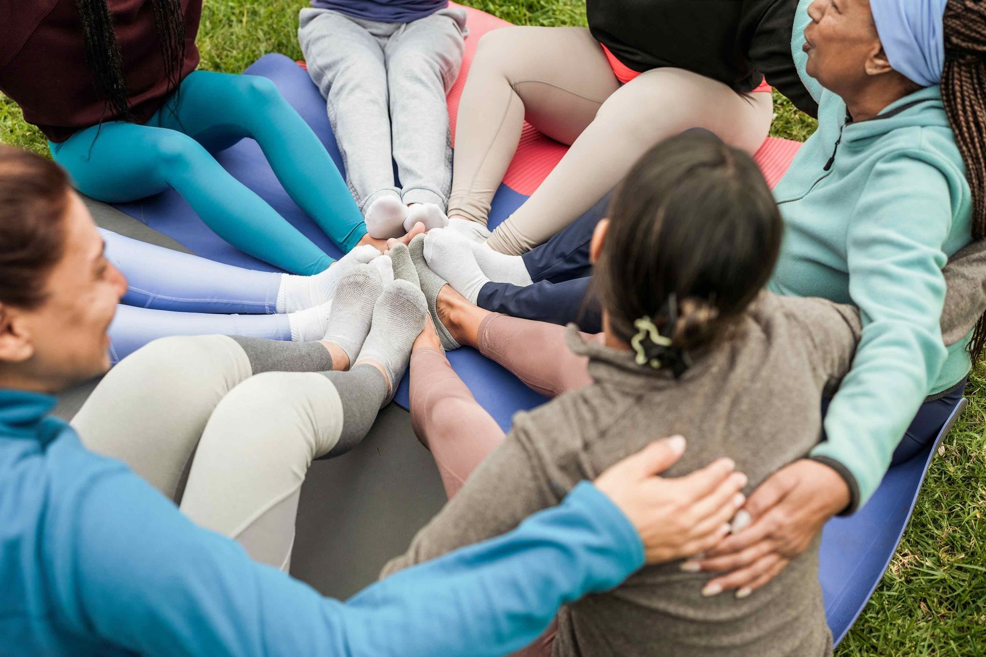 Group of people sitting in a circle, legs touching, on a mat in a grassy area. Group of people sitting in a circle, legs touching, on a mat in a grassy area.