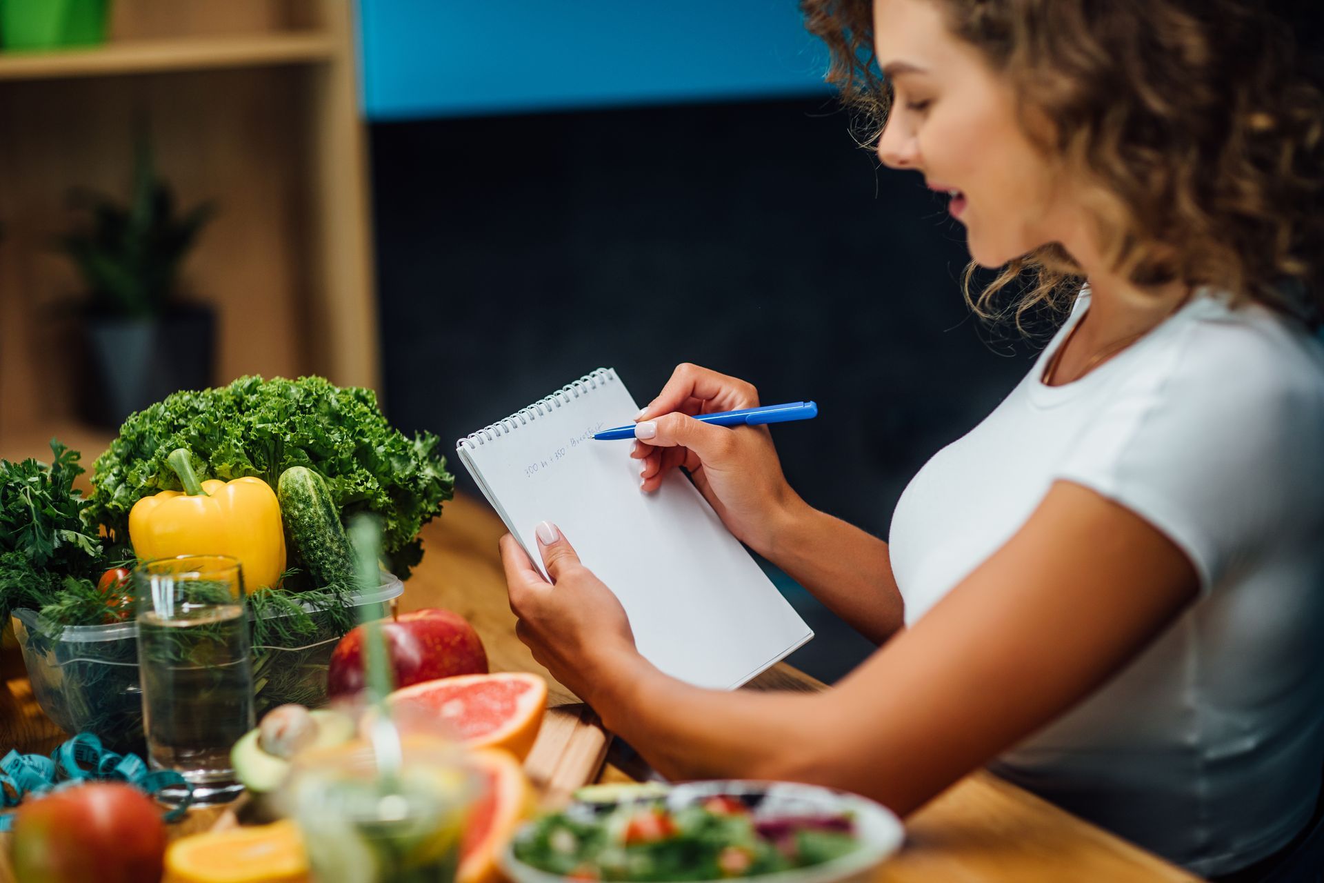 Woman writing in a notepad, surrounded by fresh fruits and vegetables.