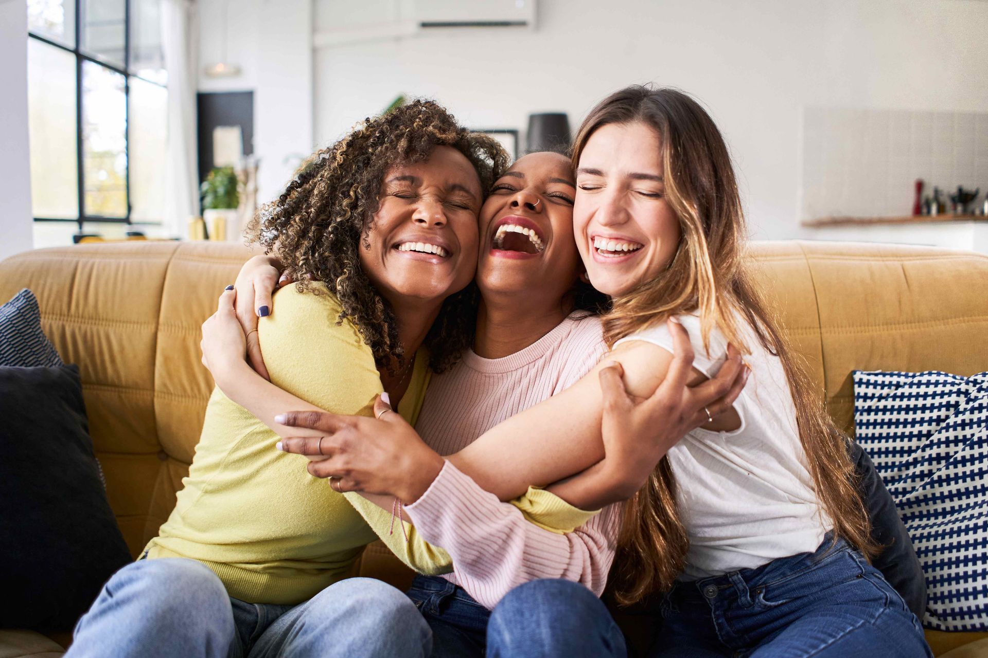 Three friends laughing and hugging on a yellow couch in a living room. Three friends laughing and hugging on a yellow couch in a living room.